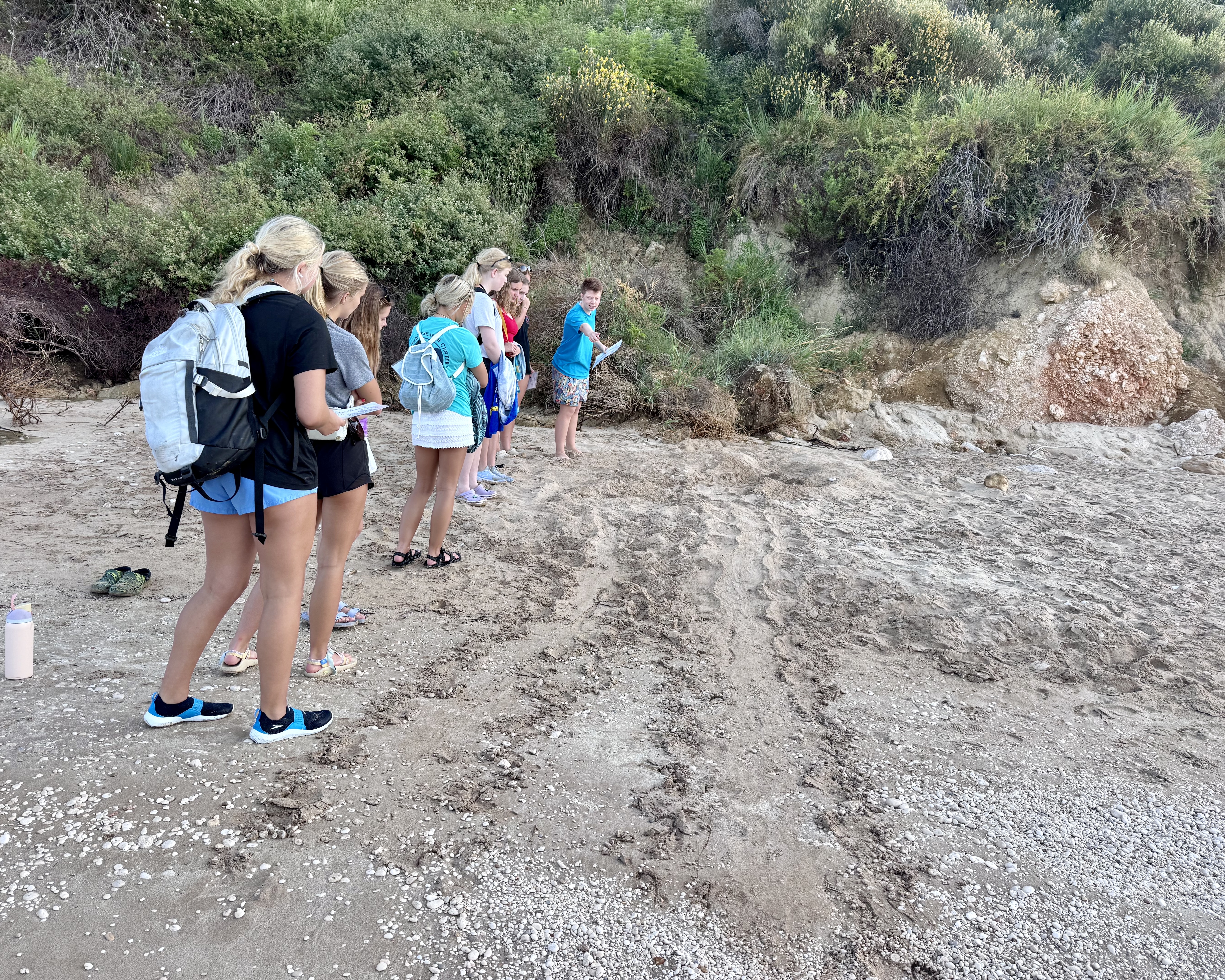 High school students in life jackets and snorkel gear wading into clear shallow water on a Kefalonia beach
