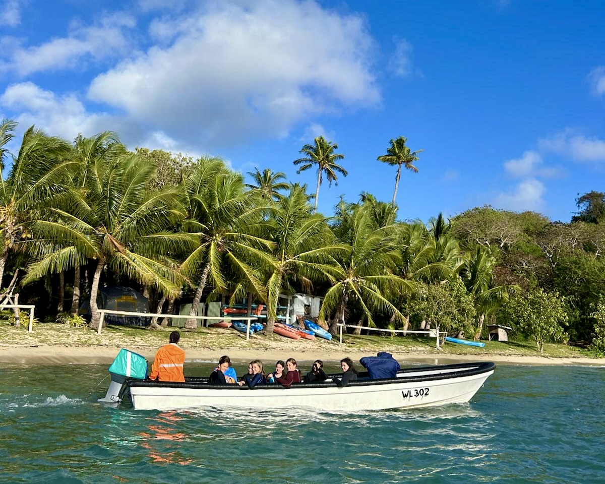 High school students riding a boat past a palm-lined beach with kayaks on the shore in the Yasawa Islands, Fiji
