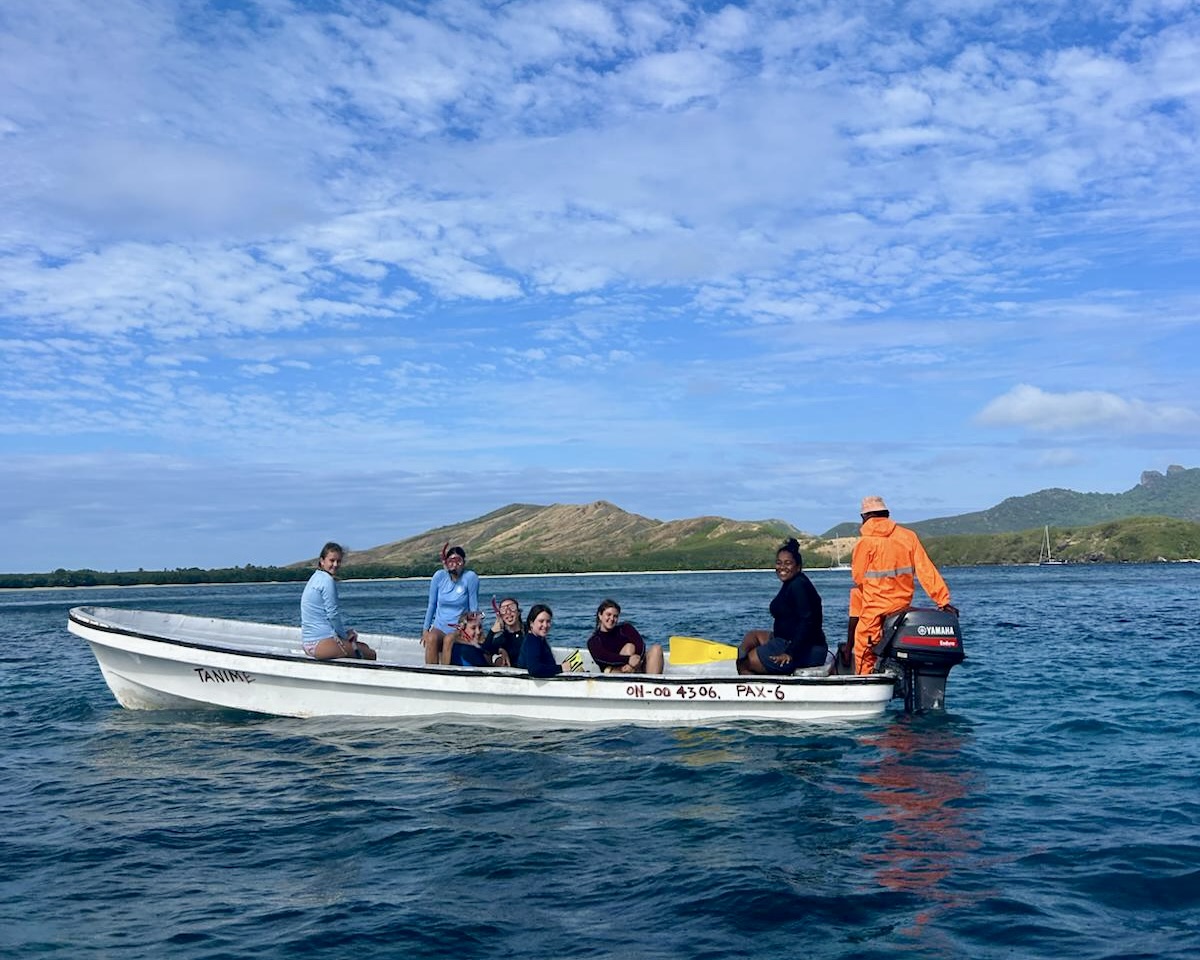 Group of high school summer program students in a boat on calm water with green island hills behind in Fiji
