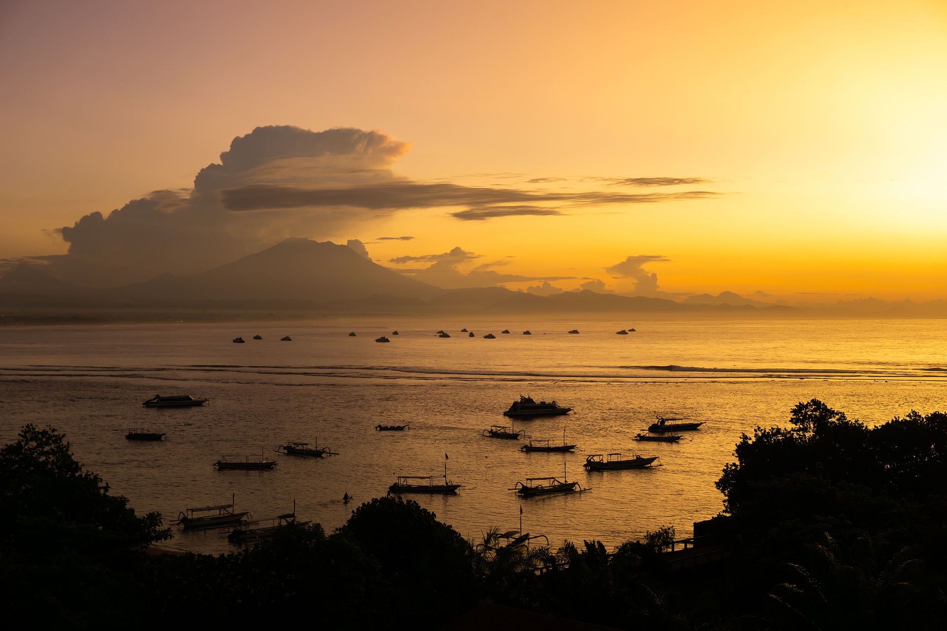Fishing boats dot a bay under an orange sunset with mountains in the distance