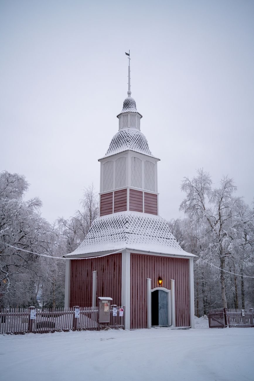 A snow-covered wooden church stands against a grey winter sky in a small northern town.