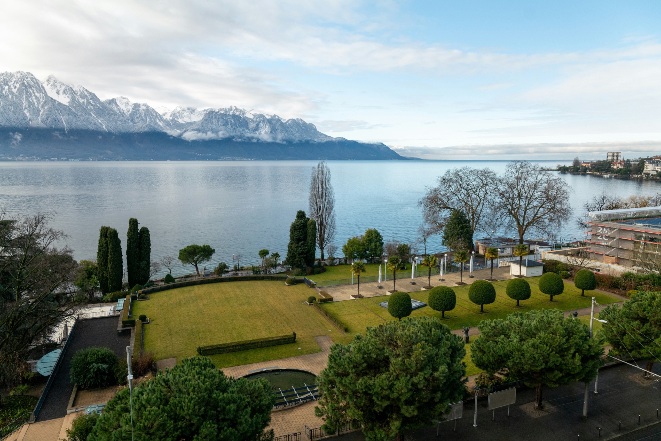 A formal lakeside garden of clipped trees and cypresses runs down to still water, with a long range of snow-covered peaks along the far shore.