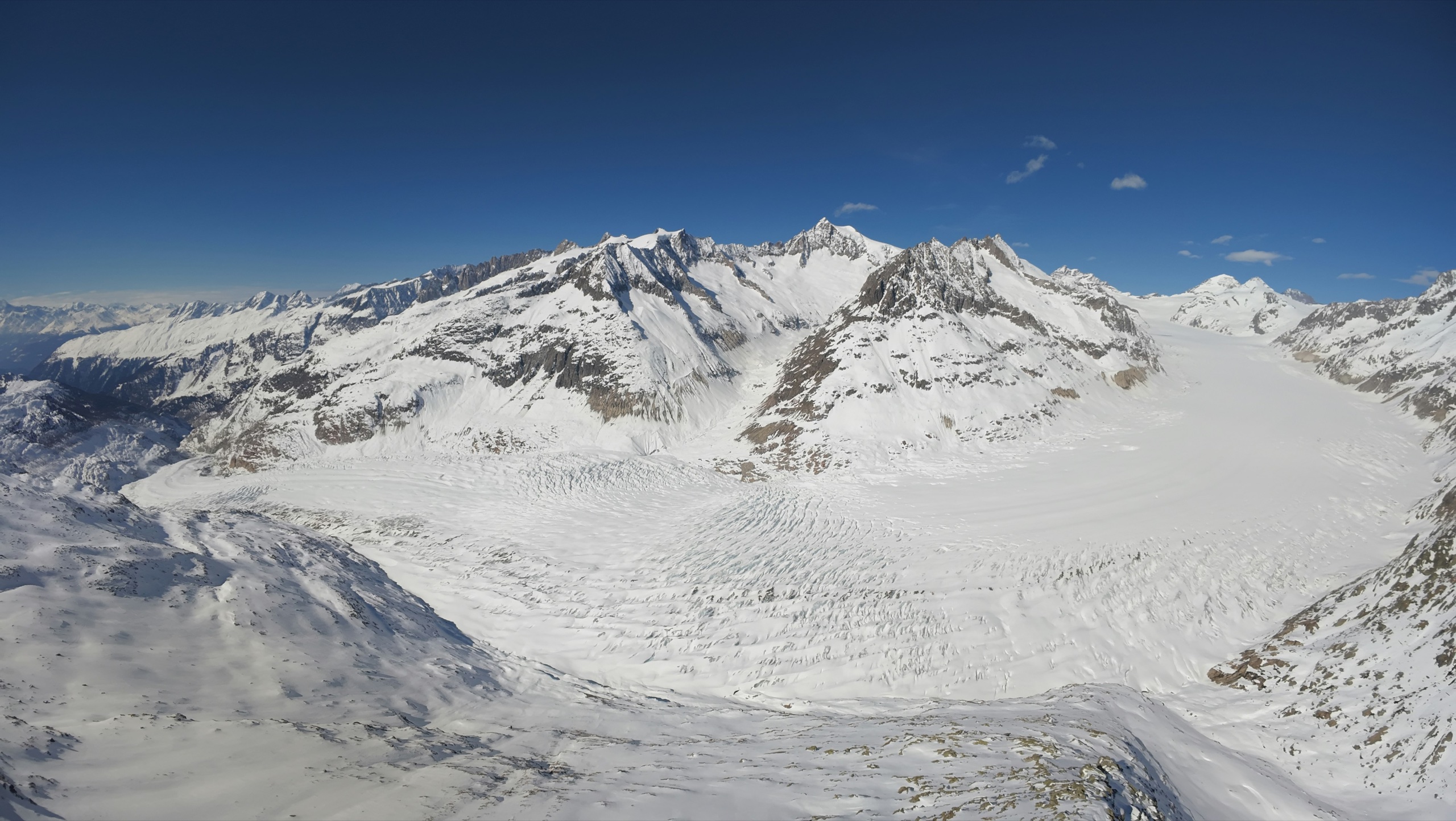 A vast glacier curves between snow-covered peaks, its surface creased with crevasses, under a deep blue sky.
