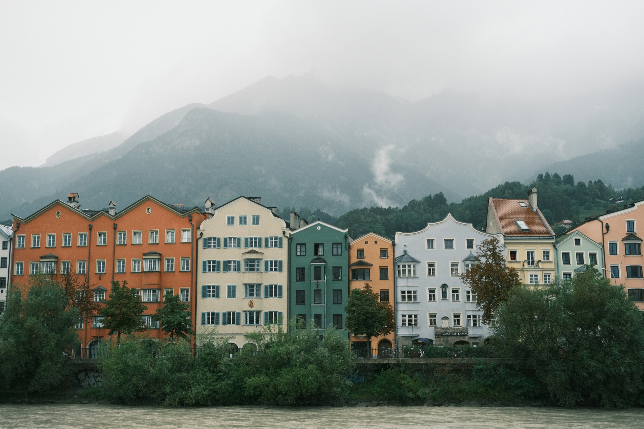 A row of tall houses in orange, cream, green, and pink lines a river bank, with a forested mountain wall rising into mist behind them.