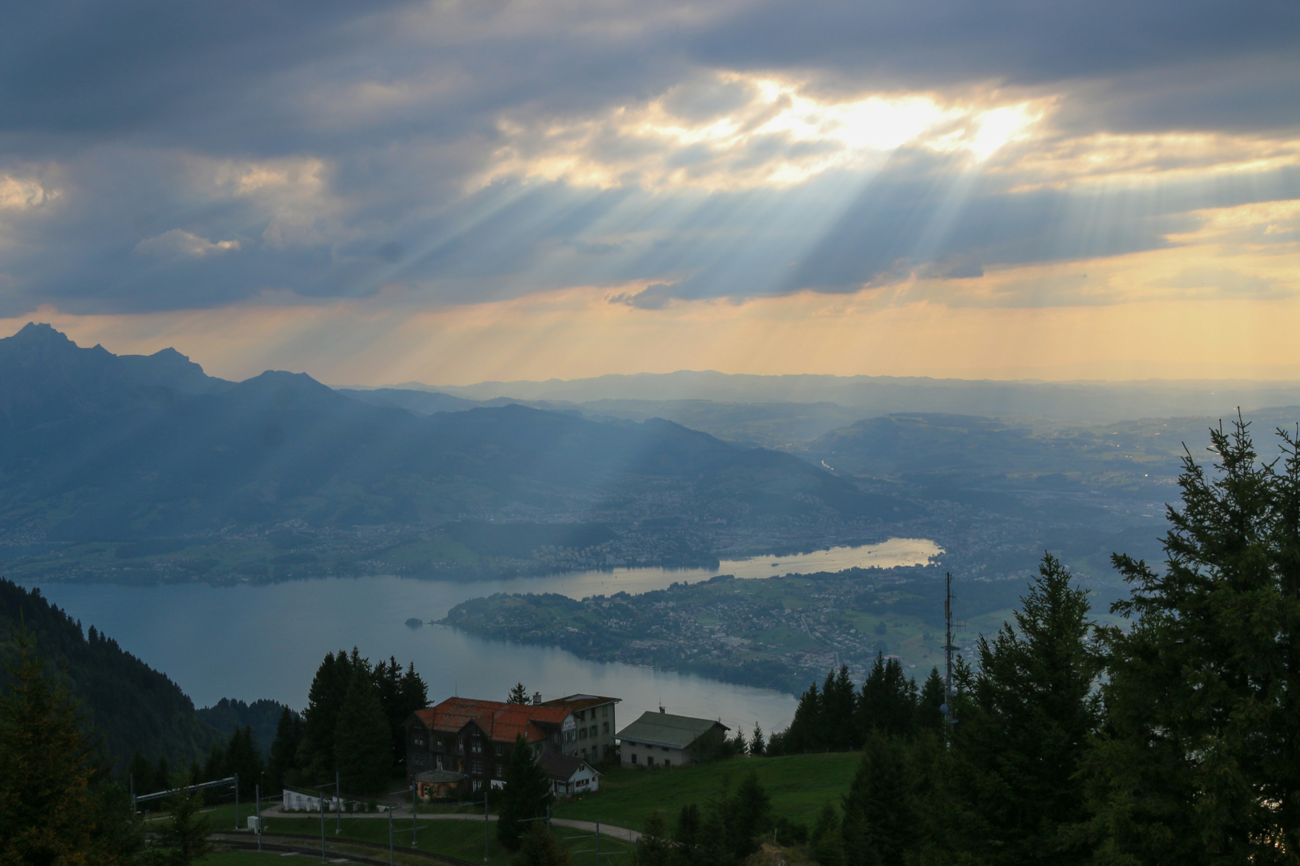Sunbeams break through cloud over a long lake far below a wooded ridge, with a hotel and a rack railway line on the slope in the foreground.