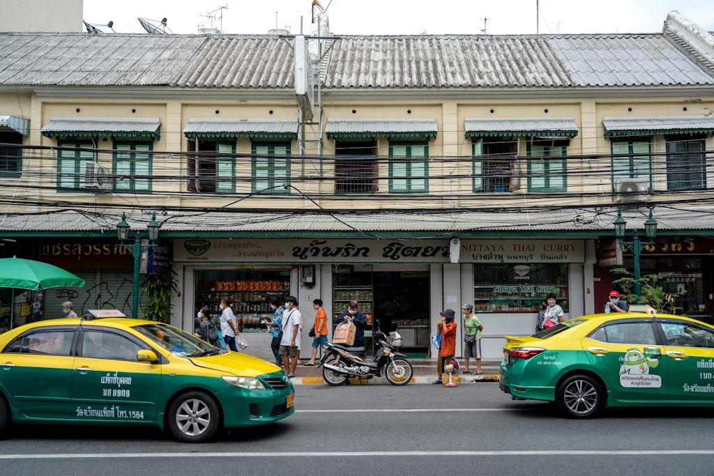 The picture features two green and yellow taxis on a street in Bangkok. Their bright colors stand out against the backdrop of a building. Passengers are in that area.