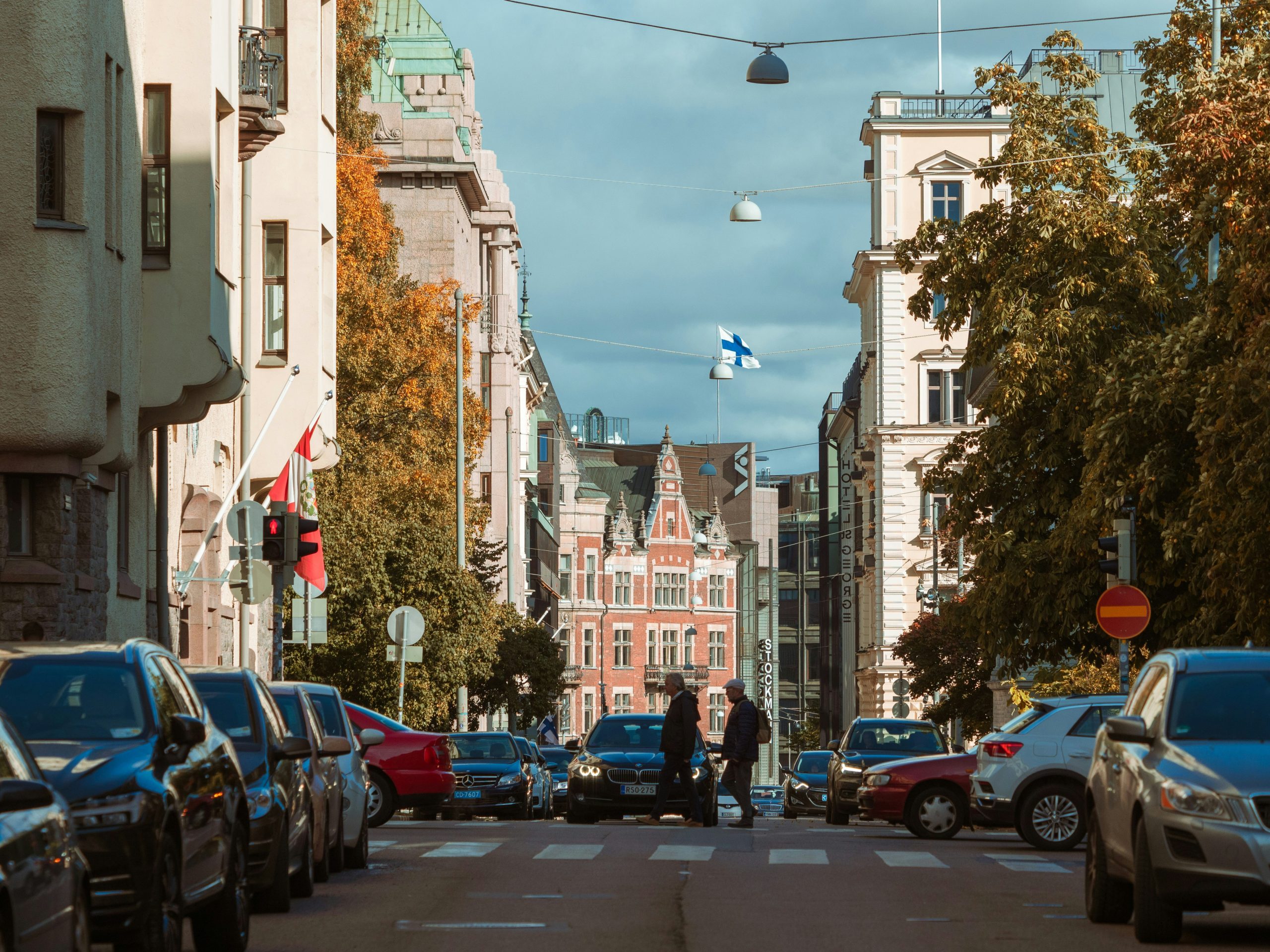 A Finnish flag flies over a Helsinki street in autumn, red-brick and pale stone facades rising past parked cars and yellowing trees.