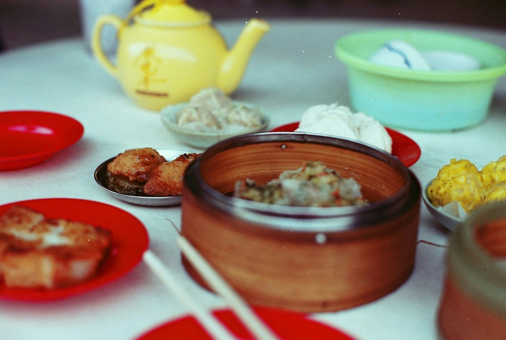 A spread of dim sum dishes, including dumplings, buns, and fried treats, served with a yellow teapot and red plates on a table.