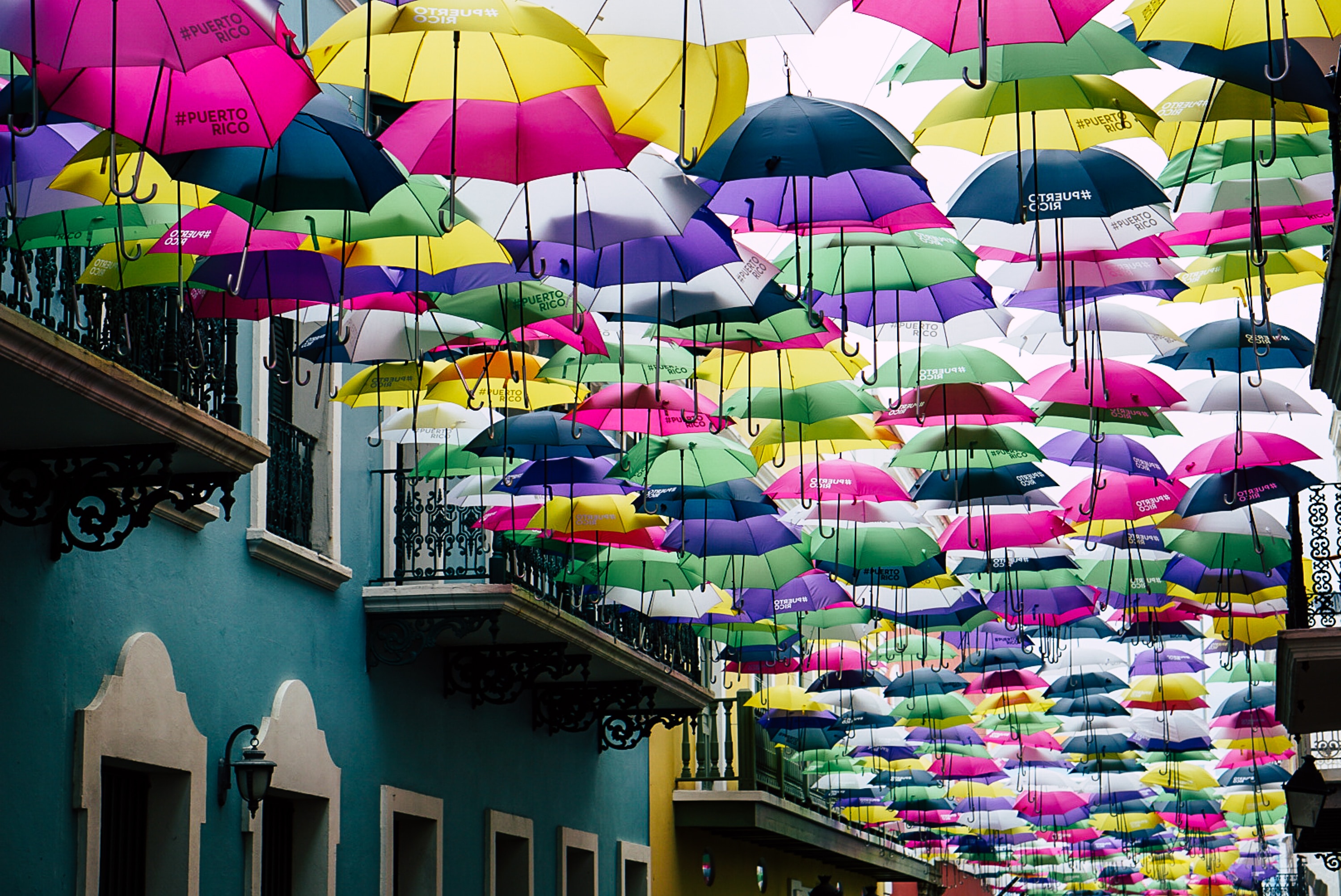 A school trip stops beneath colorful umbrellas hanging above a narrow street between pastel buildings