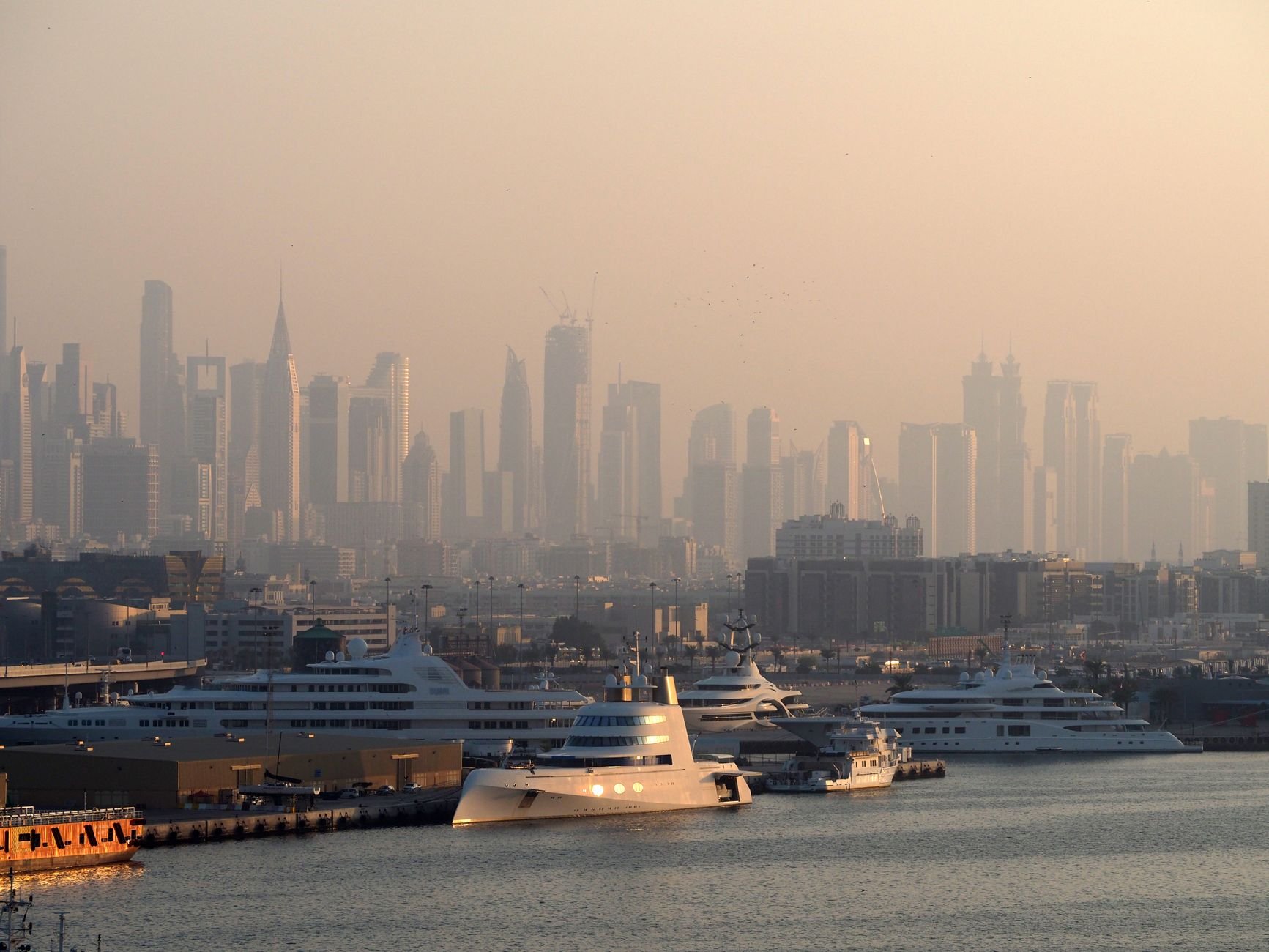 A city skyline across the harbor at dusk, with private yachts moored in the foreground and the towers softened by haze.