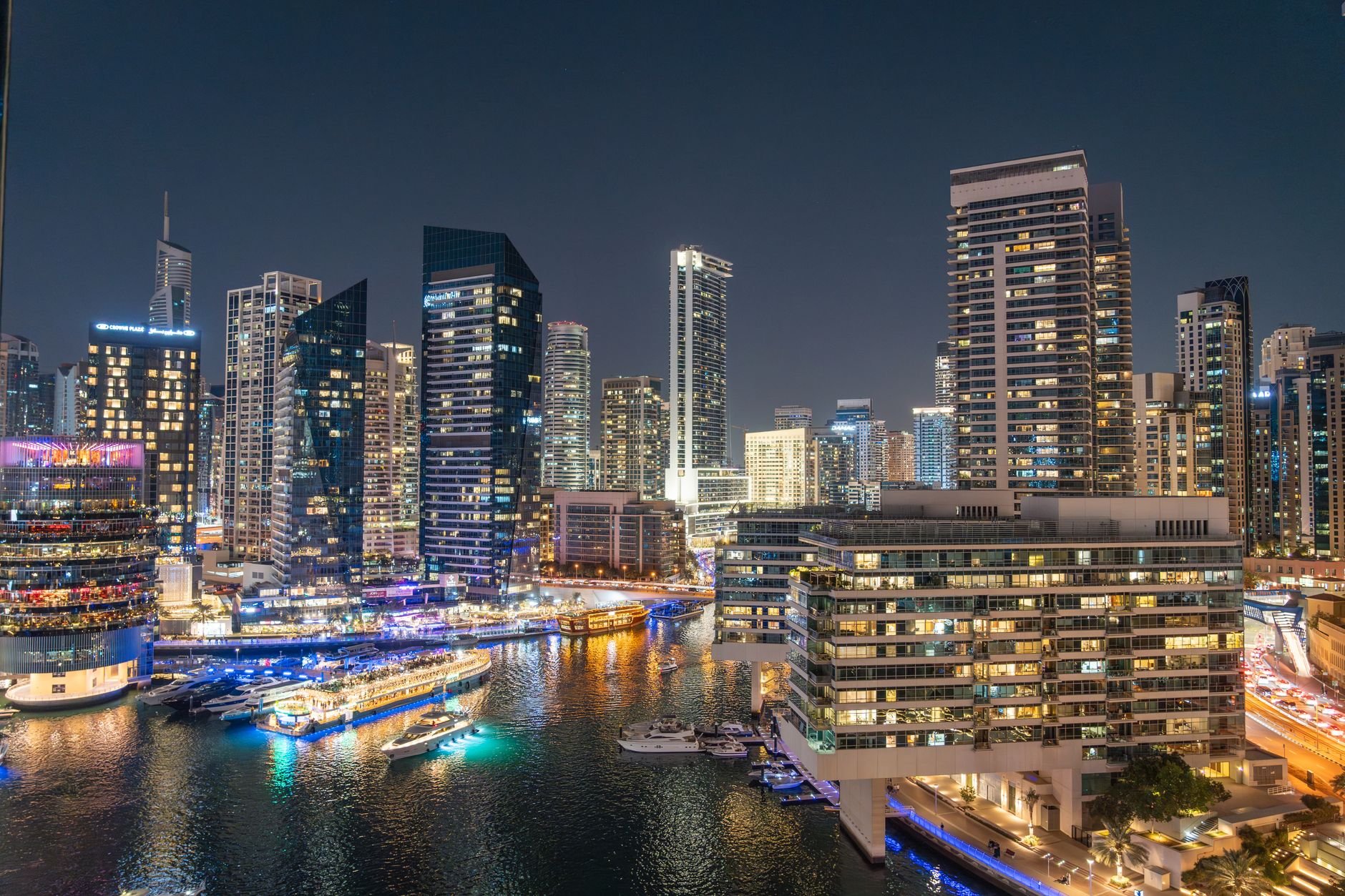 A marina illuminated at night, with towers reflected in the water and boats moored along the promenade.