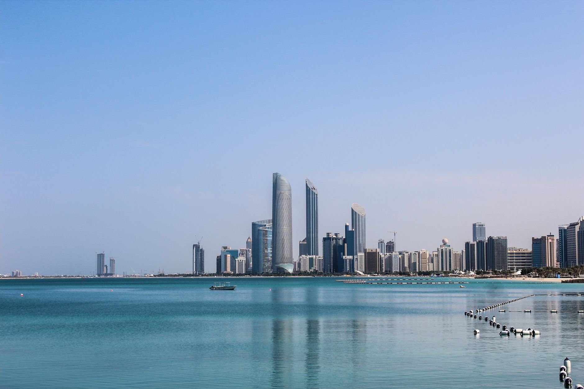 A city skyline across a calm turquoise bay, with a small boat on the water and a roped swimming area in the foreground.