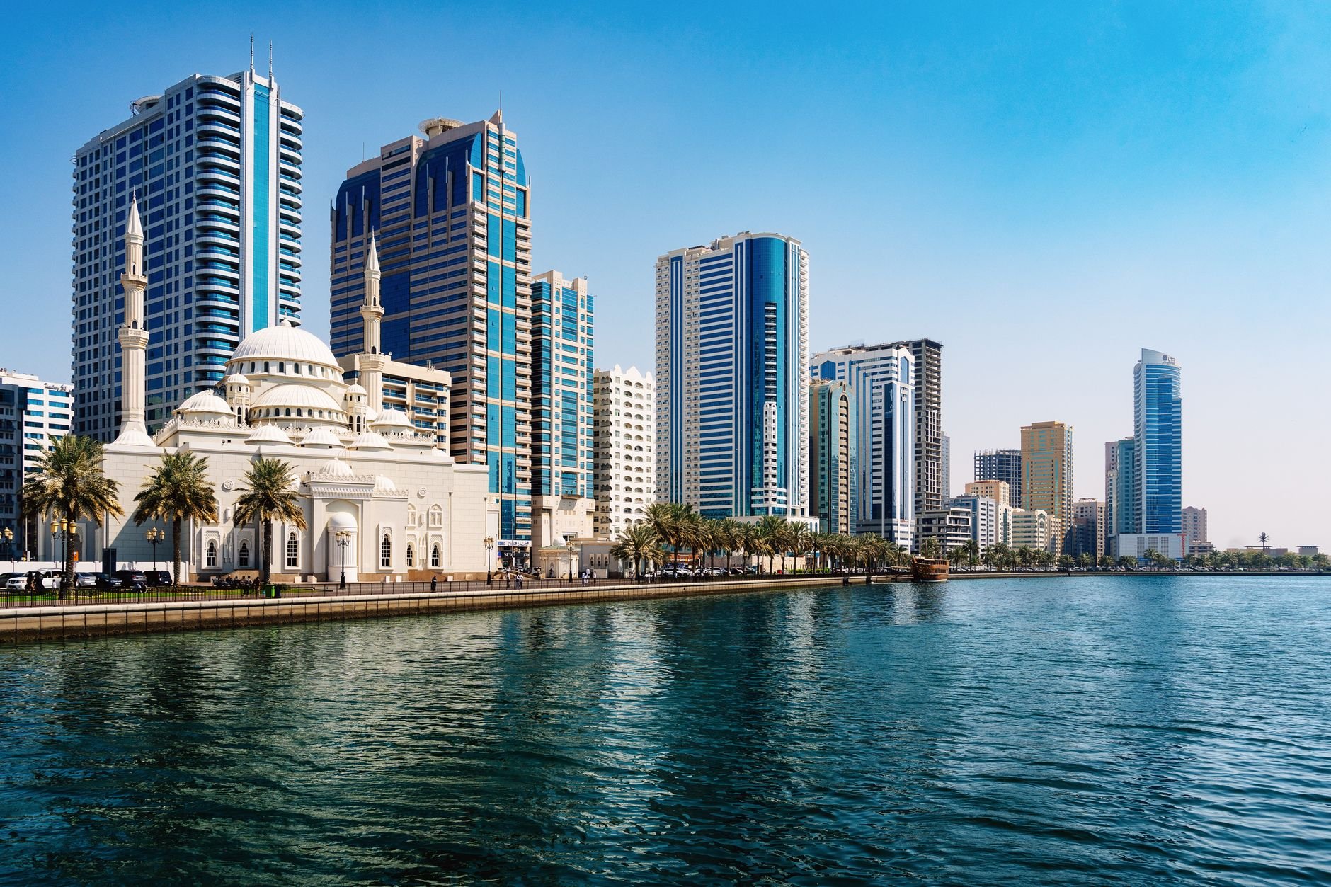 A white domed mosque with twin minarets on a waterfront corniche, framed by palm trees and towers.