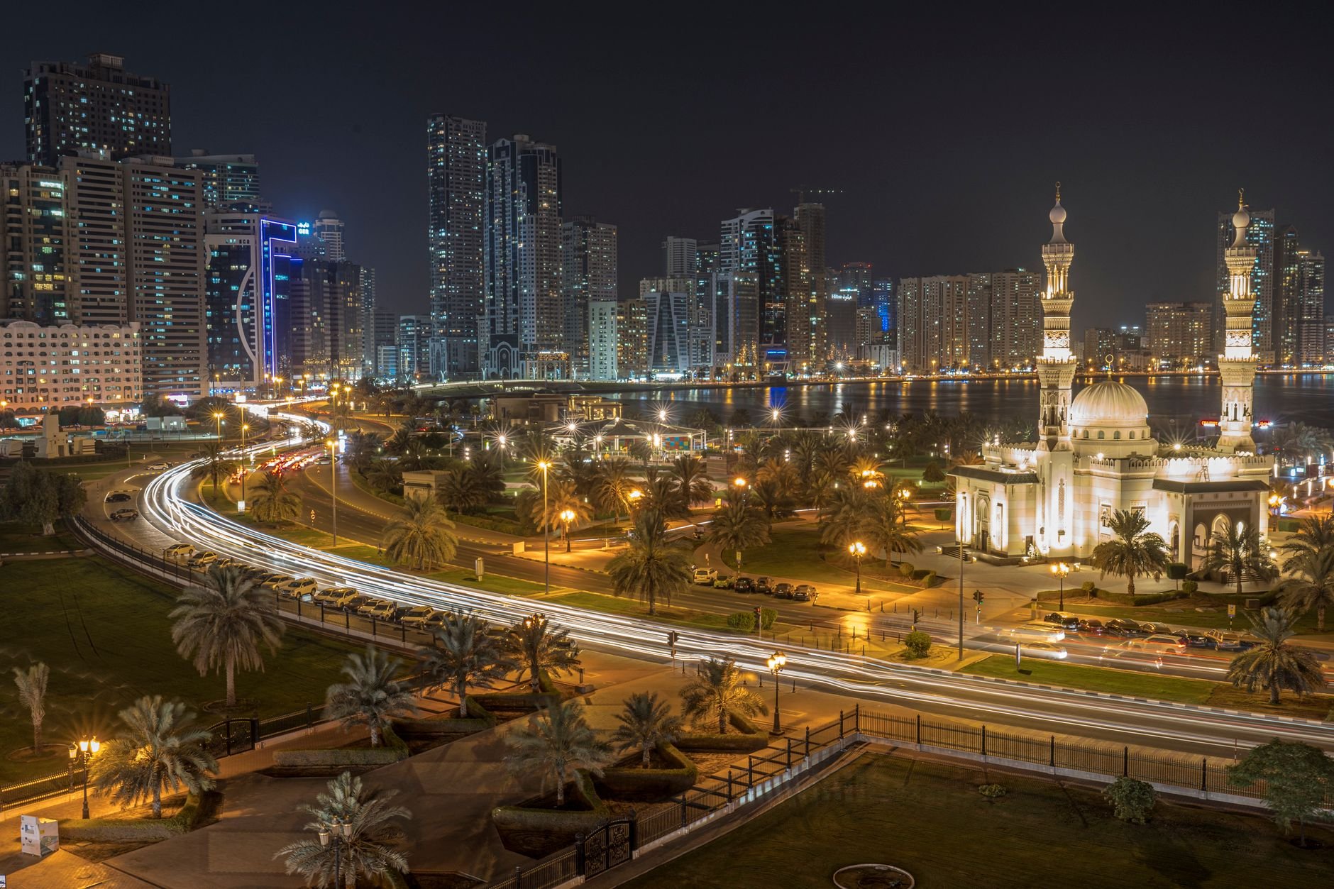 A skyline and an illuminated mosque at night, seen across a corniche with light trails from passing traffic.