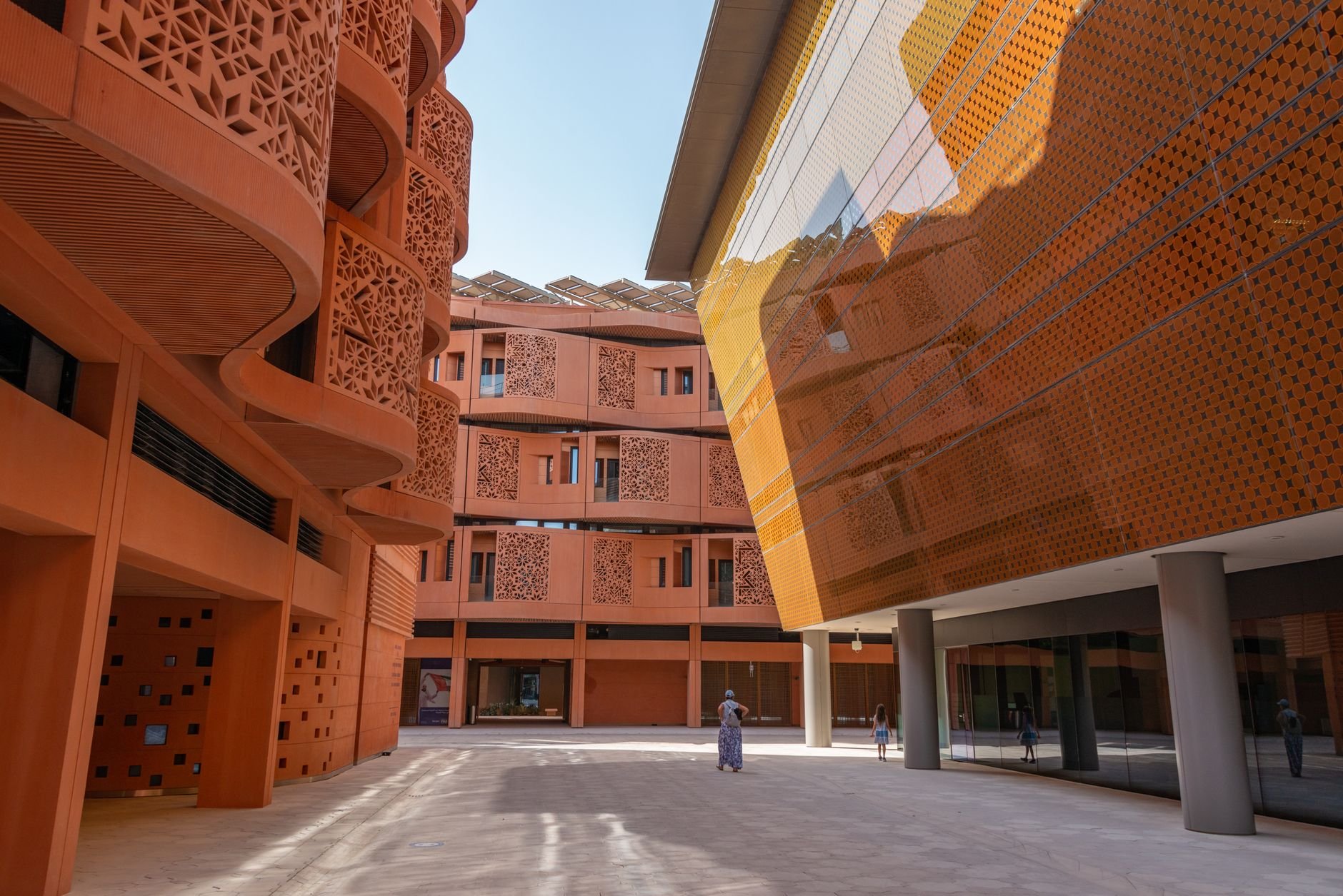 A sunlit courtyard between terracotta lattice-screened buildings, with a few visitors walking between them.