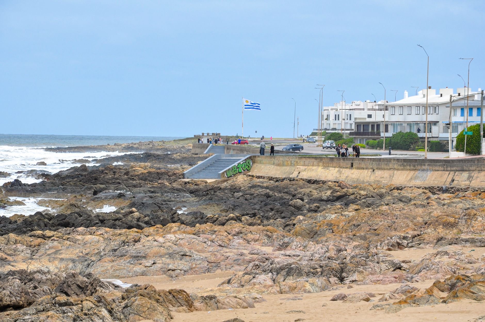 A rocky seafront promenade with a flagpole and low white buildings beyond.