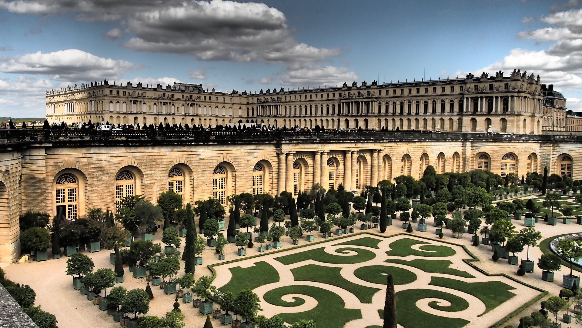 A school trip pauses among ornate garden parterres and manicured hedges lining the wings of a grand palace