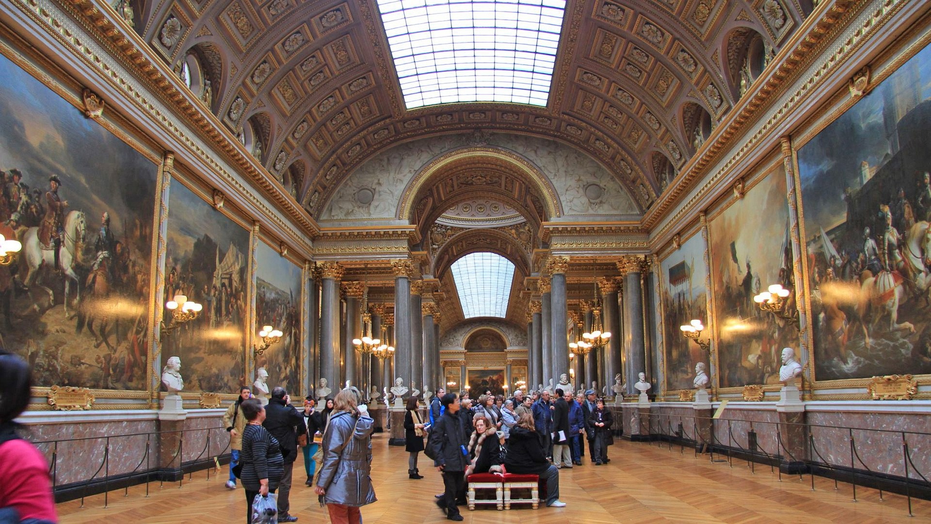 Visitors gather beneath the vaulted, painted ceiling of an ornate palace gallery lined with statues and large paintings