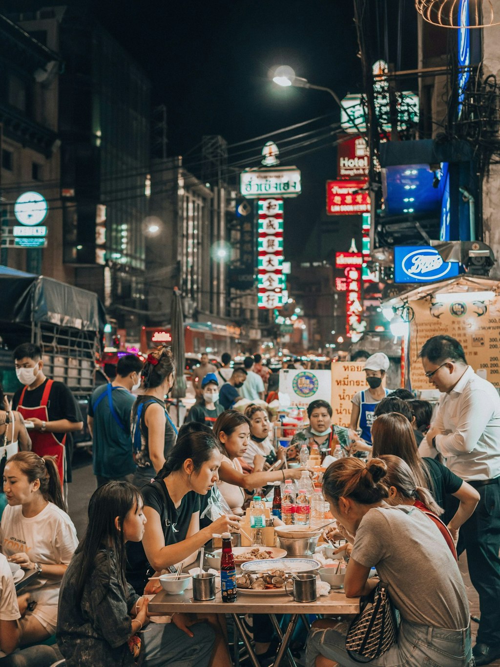 The picture shows a busy street in Bangkok's Chinatown. People are eating food at small tables, and vendors display various dishes at their stalls. Colorful signs surround the area, and the scene is full of activity.