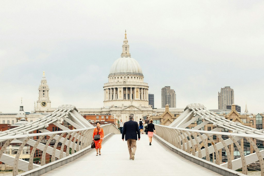 A bright view of the dome of Saint Paul's Cathedral can be seen in the background as people walk across the Millennium Bridge.