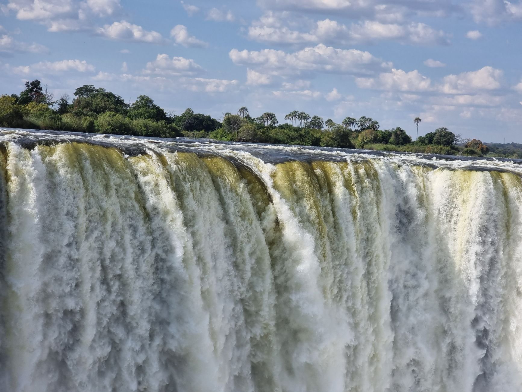 A waterfall in Zimbabwe seen close to the lip, water pouring over the edge in heavy flow with green forest beyond.
