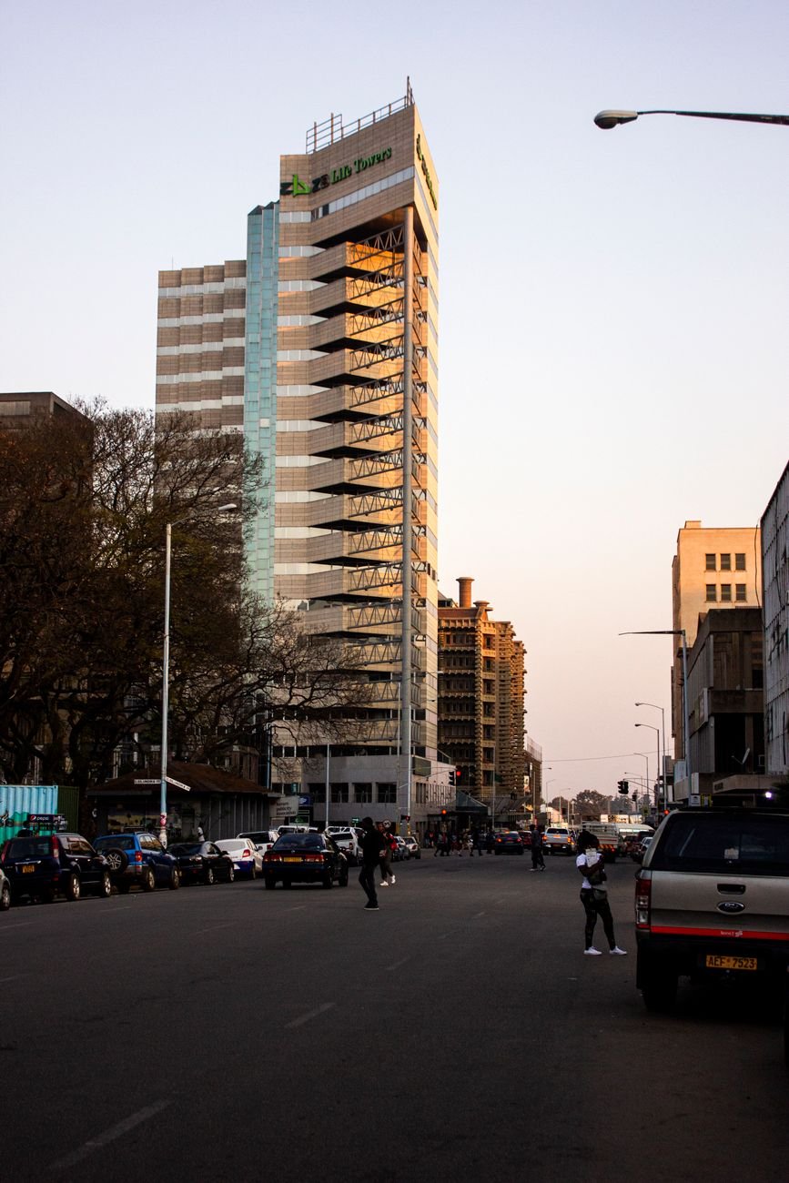 A Harare street at dusk with a tall office tower lit by the setting sun and pedestrians crossing below.