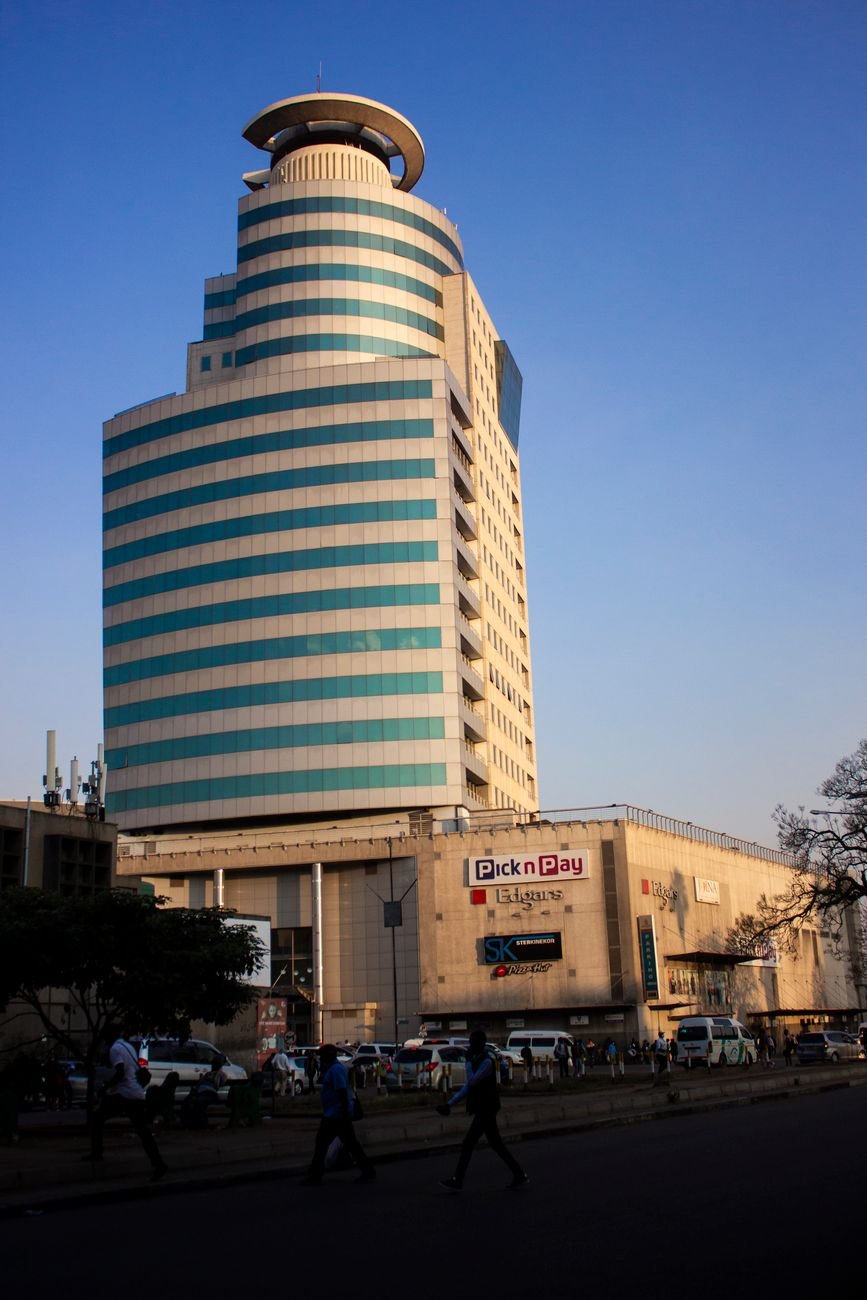 A curved glass-and-stone office tower above a shopping arcade in Harare, with pedestrians on the sidewalk.